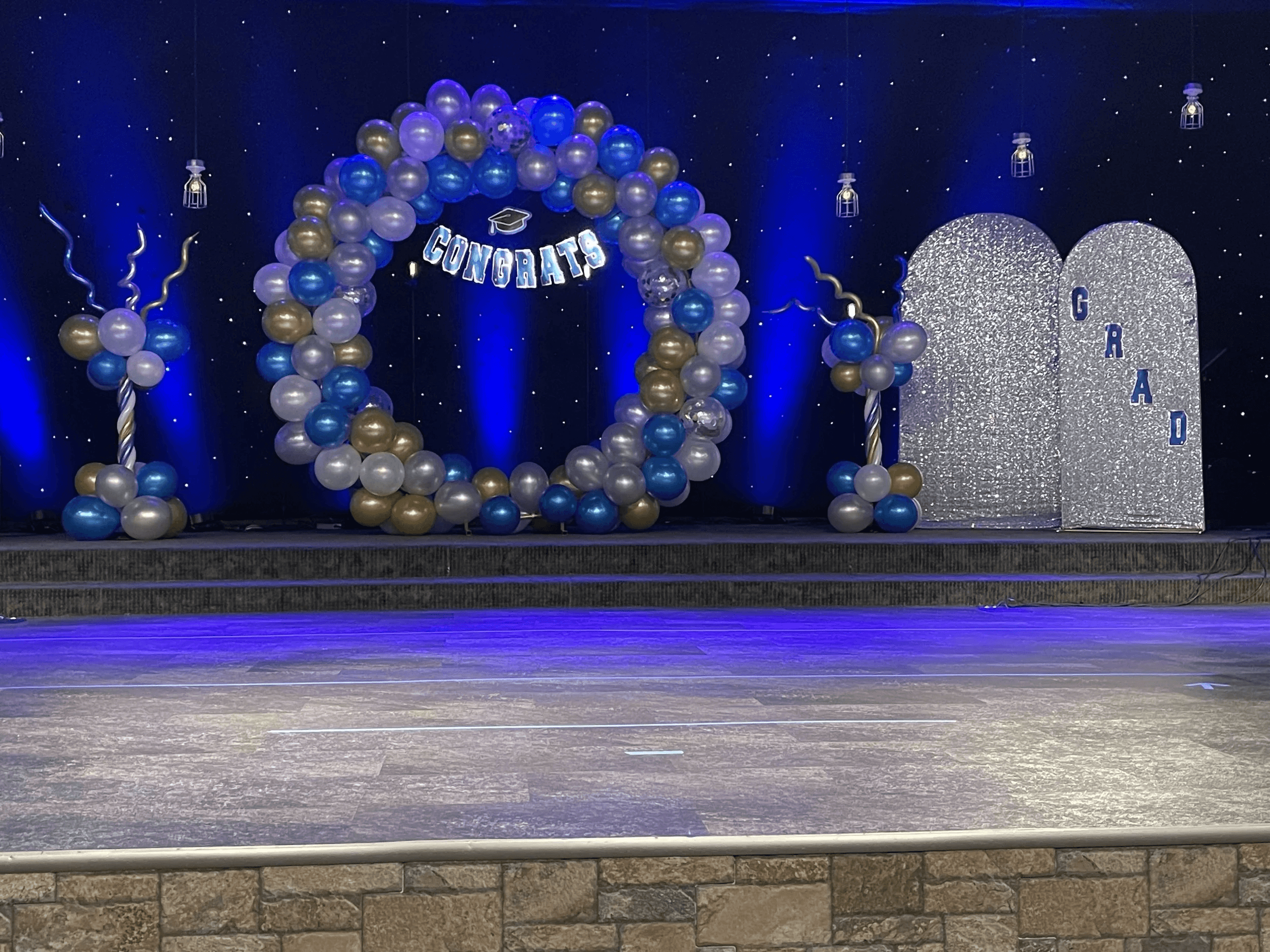 Arco de globos de graduación en azul y dorado con pancarta de felicitación en un escenario